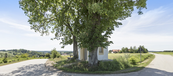 Eine Straße macht einen Bogen, in der Mitte des Bogens ist eine Wiese mit Bäumen, die eine kleine Kapelle verdecken.